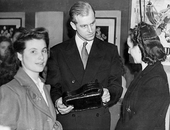 The Duke of Edinburgh examines a handbag made by girls of the leatherwork and millinery club during a visit to the Dockland Settlement. 1947.